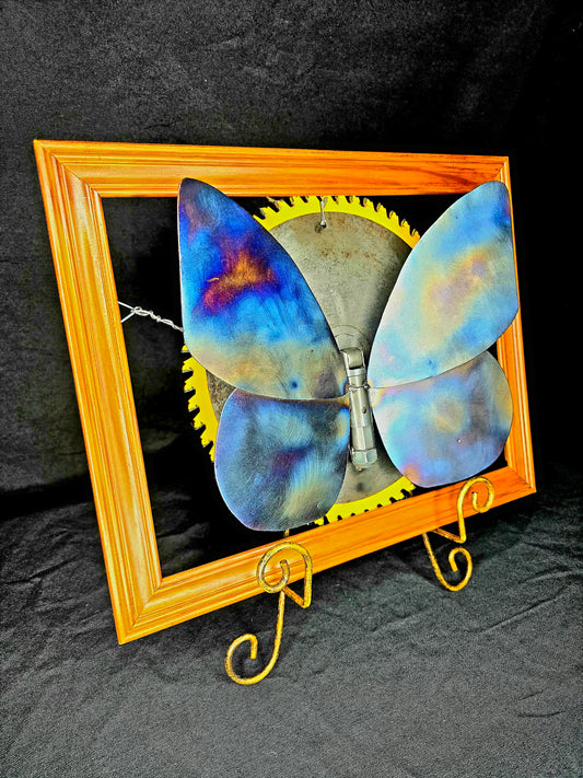 The Transformation: From Cocoon to Flight is a metal butterfly sculpture by Enrique Del Angel displayed within a wood frame.