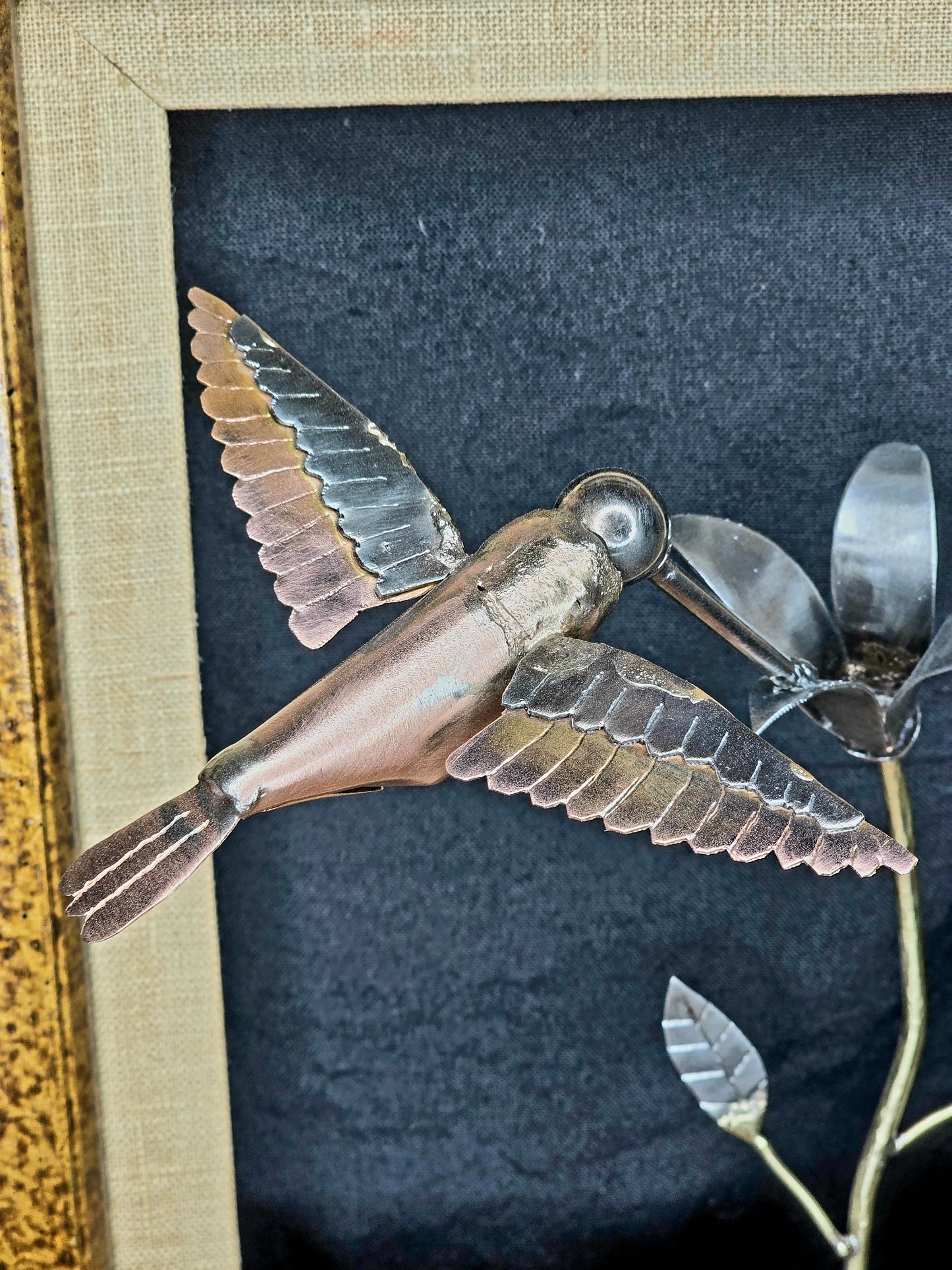 Close-up of framed metal bird sculpture with copper body, silver wings, and flower stem