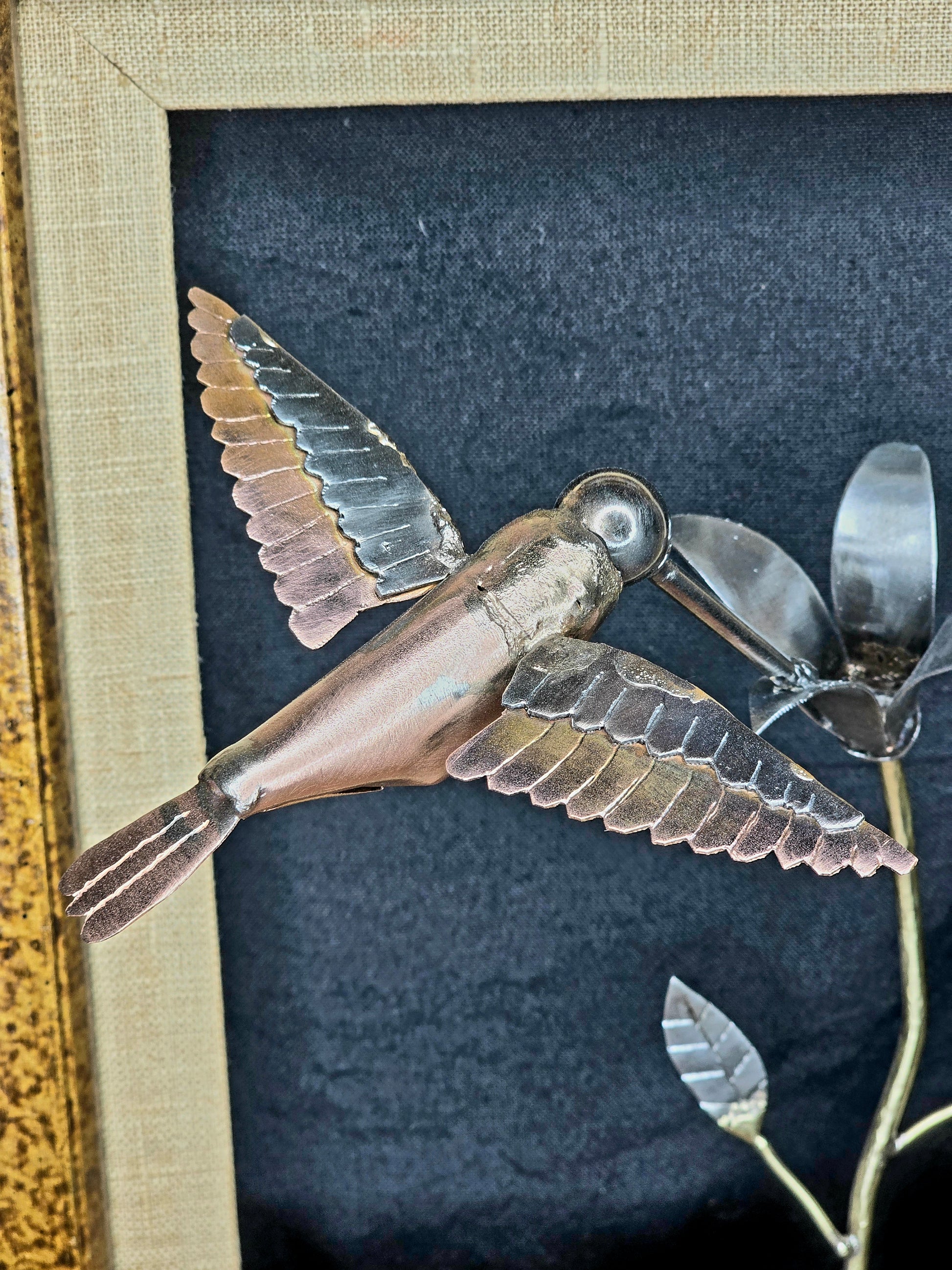 Close-up of framed metal bird sculpture with copper body, silver wings, and flower stem