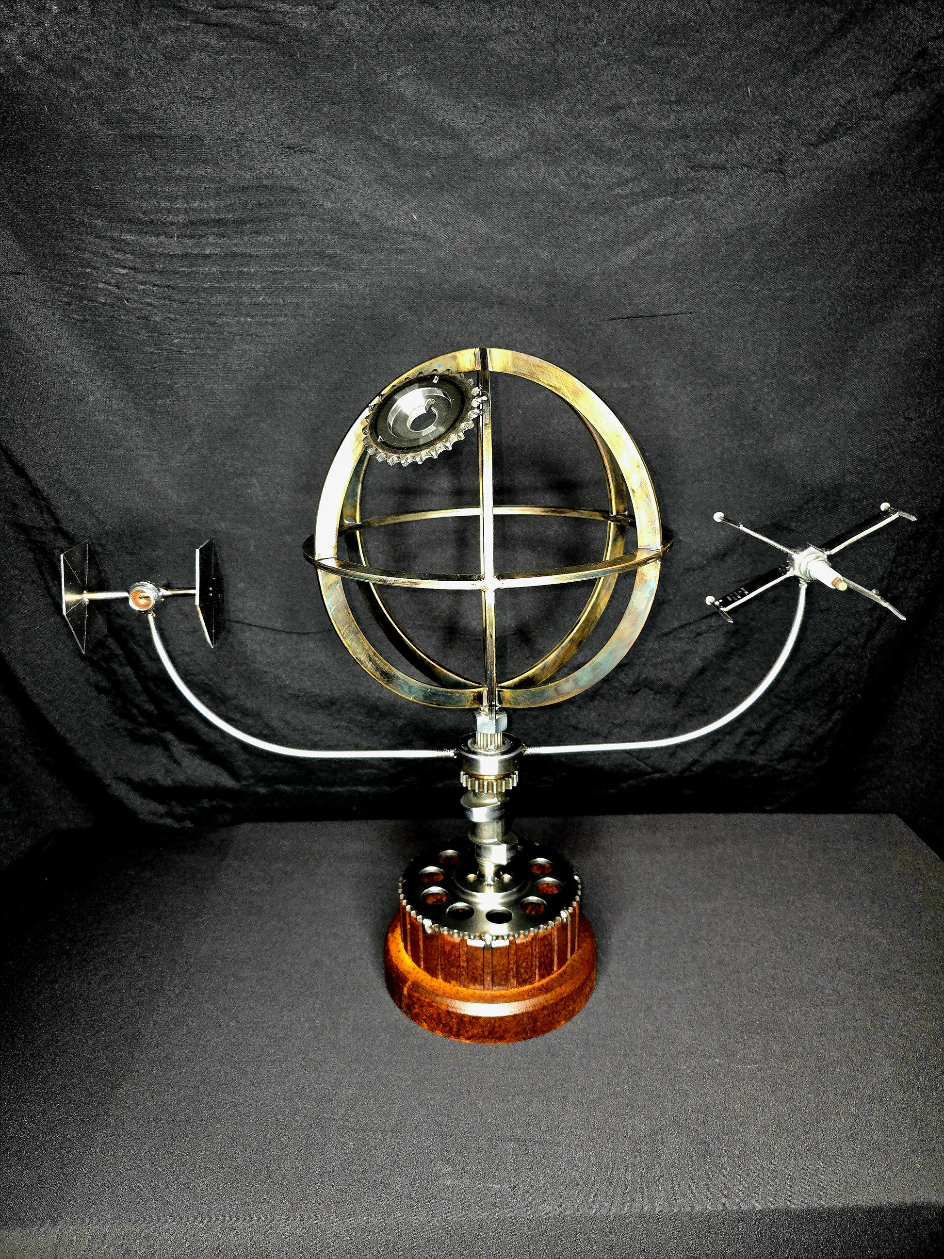 The Death Star kinetic metal sculpture showing a brass globe death star model with curved mechanical arms holding a TIE Fighter and X-Wing starship over a wood and gear base.