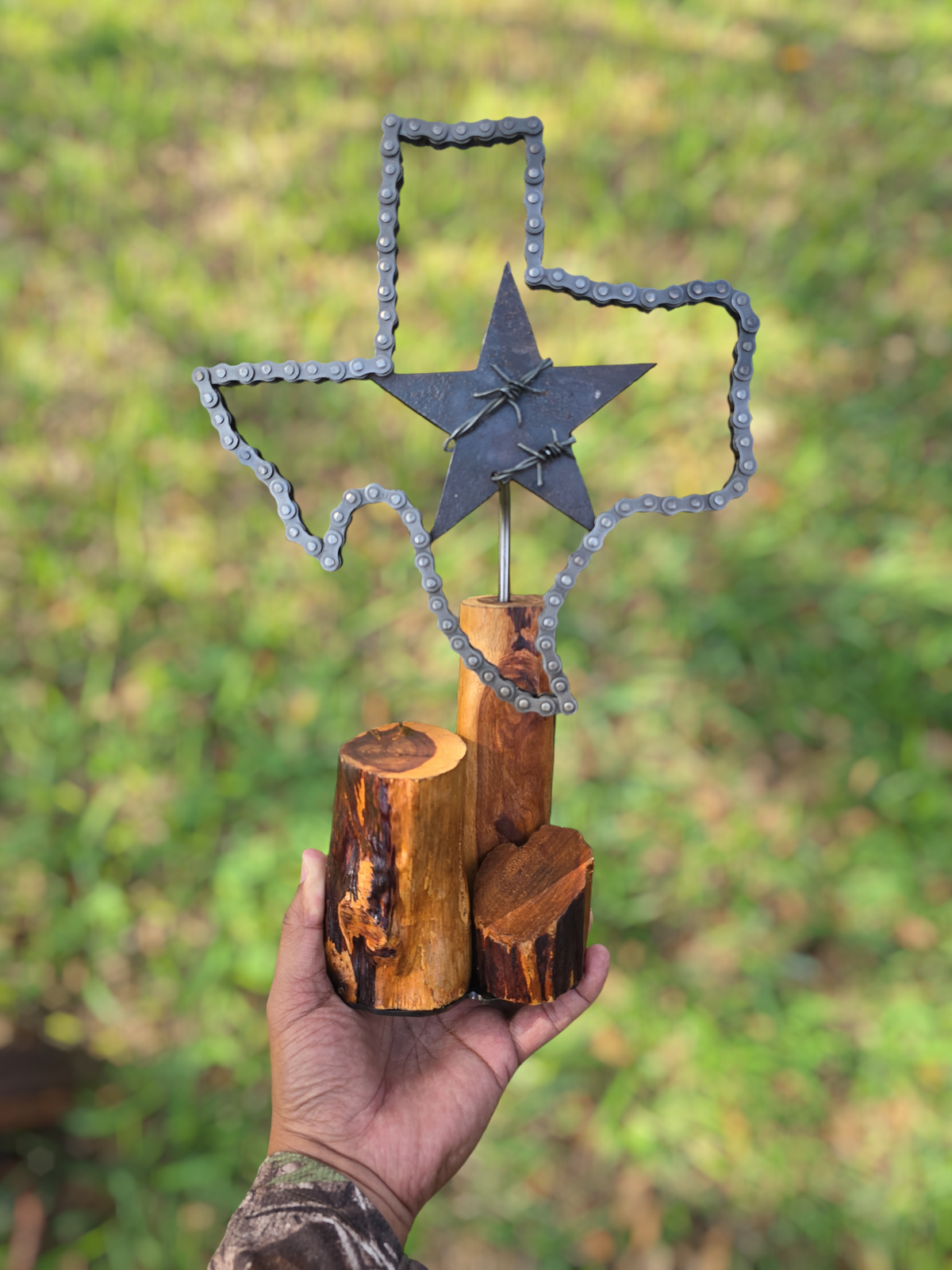 Texas metal sculpture by Enrique Del Angel. Features a Texas outline made of automotive chain, a barbed wire star, brass shell casings, and an Ebano wood base.