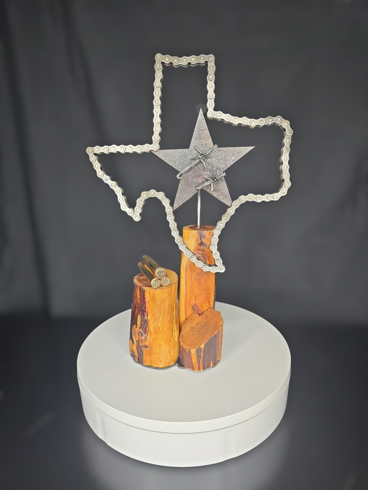 Texas metal art sculpture 'Where the Brass Falls, the Lone Star Rises' by Enrique Del Angel. Features a Texas outline made of automotive chain, a barbed wire star, and brass shell casings on an Ebano wood base.